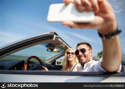 road trip, leisure, couple, technology and people concept - happy man and woman driving in cabriolet car and taking selfie with smartphone