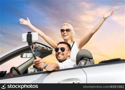 road trip, technology and travel concept - happy couple driving in convertible car and taking picture by smartphone on selfie stick over sky background. couple in car taking selfie by smartphone over sky. couple in car taking selfie by smartphone over sky