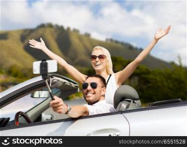 road trip, technology and travel concept - happy couple driving in convertible car and taking picture by smartphone on selfie stick. happy couple in car taking selfie by smartphone. happy couple in car taking selfie by smartphone