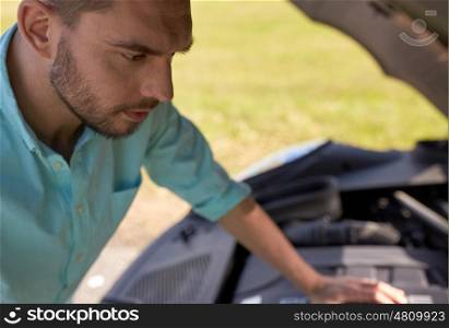 road trip, transport, travel and people concept - close up of young man with open hood of broken car at countryside