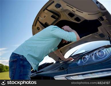 road trip, transport, travel and people concept - close up of young man with open hood of broken car at countryside