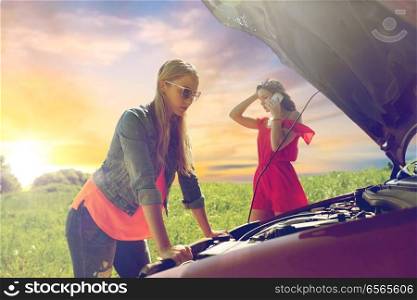 road trip, transport, travel and people concept - young women with open hood of broken car at countryside calling on smartphone to evacuation service over evening sky background. women with open hood of broken car at countryside