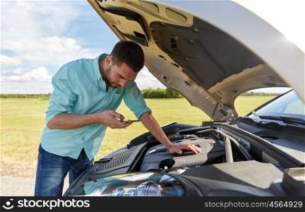 road trip, transport, travel, technology and people concept - young man with smartphone and open hood of broken car at countryside