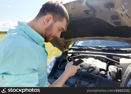 road trip, transport, travel, technology and people concept - young man with smartphone and open hood of broken car at countryside