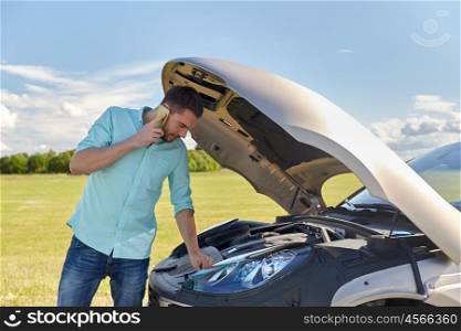 road trip, transport, travel, technology and people concept - young man with smartphone and open hood of broken car at countryside calling to evacuation service