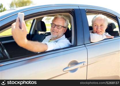 road trip, travel and old people concept - happy senior couple driving in car and taking selfie by smartphone . senior couple in car taking smartphone selfie
