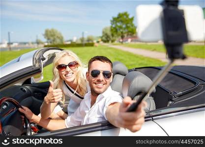 road trip, travel, couple, technology and people concept - happy man and woman driving in cabriolet car and taking picture with smartphone on selfie stick