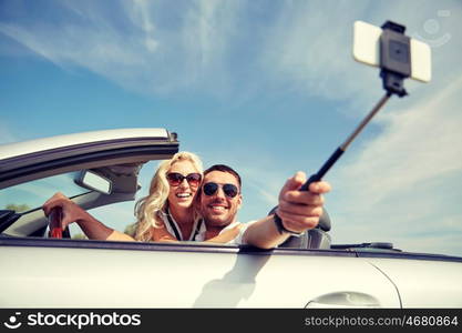 road trip, travel, couple, technology and people concept - happy man and woman driving in cabriolet car and taking picture with smartphone on selfie stick