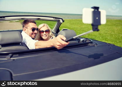 road trip, travel, couple, technology and people concept - happy man and woman driving in cabriolet car and taking picture with smartphone on selfie stick