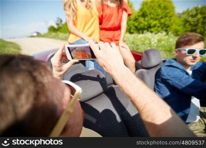road trip, travel, technology and people concept - happy friends driving in cabriolet and photographing by smartphone outdoors