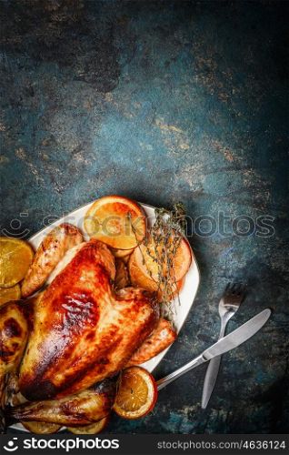 Roast chicken on platter and roasted orange slices served with fork and knife on rustic background, top view
