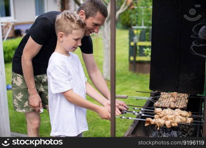 roast meat, food on fire, coals, fire, marinade, nature, fun company, family day, white t-shirt, cooking, cooking, cooking, fatherhood. Cheerful dad and son roast meat on the grill