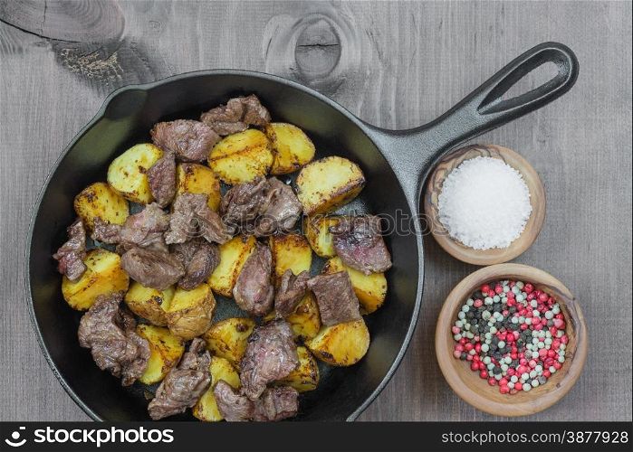 Roast meat with potato in a cast iron skillet and two wooden bowls with salt and multicolored pepper on a wooden table