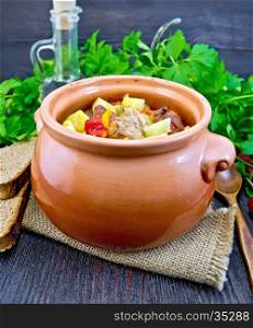 Roast with chicken, potatoes, squash and sweet peppers in a clay pot on a napkin of burlap, spoon, parsley on a dark wooden board