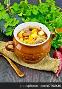 Roast with chicken, potatoes, squash, and sweet peppers in a portion clay pot on a napkin of burlap, spoon, parsley on a dark wooden board