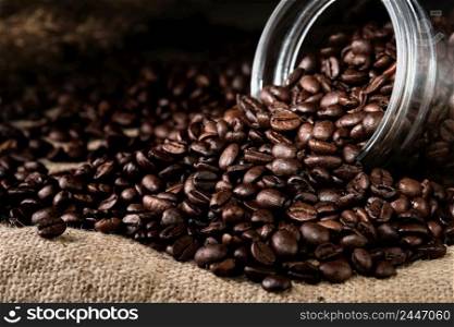 Roasted aromatic coffee beans are scattered from a jar on burlap. Close-up, selective focus. Copy space banner.