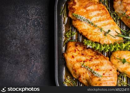 Roasted chicken breast in grill pan with fresh herbs on dark rustic background, top view, close up