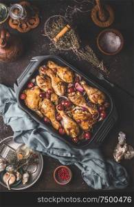 Roasted chicken legs drumsticks with red onion and grapes in cast iron casserole pan on dark rustic background with kitchen utensils herbs and spaces, top view. Flat lay