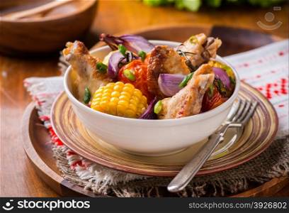 Roasted chicken with vegetables in bowl. Chicken wings, tomato, corn and onion. Green background, selective focus, rustic