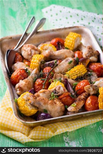 Roasted chicken with vegetables in metal baking tray. Chicken wings, tomato, corn and onion. Green background, selective focus