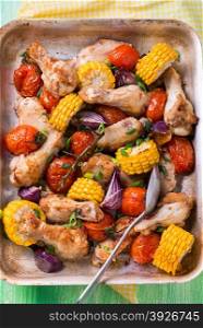 Roasted chicken with vegetables in metal baking tray. Chicken wings, tomato, corn and onion. Green background, top view