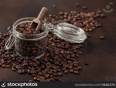 Roasted coffee beans in glass jar with wooden scoop on brown background. Macro