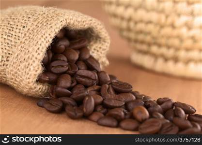 Roasted coffee beans in jute bag on wood with woven basket in the back (Selective Focus, Focus on the coffee beans turned to the front in the middle of the image)