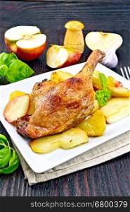 Roasted duck leg with apple, potatoes in a dish on towel, basil, garlic and fork on the background of wooden boards