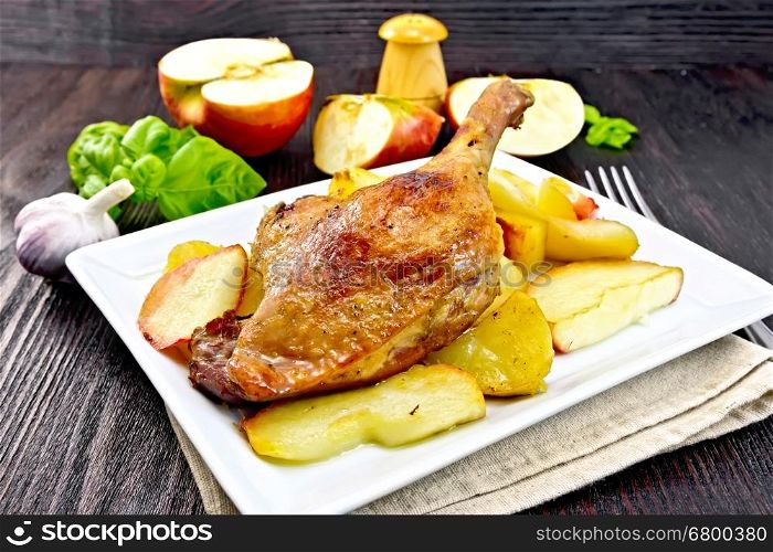 Roasted duck leg with apple, potatoes in a plate on a napkin, basil, garlic and fork on a dark wooden board