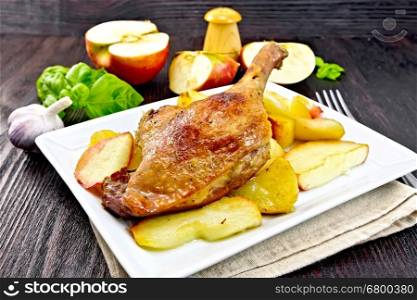 Roasted duck leg with apple, potatoes in a plate on a napkin, basil, garlic and fork on a dark wooden board