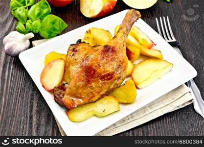 Roasted duck leg with apple, potatoes in a plate on a napkin, green basil, garlic and fork on the background of wooden boards