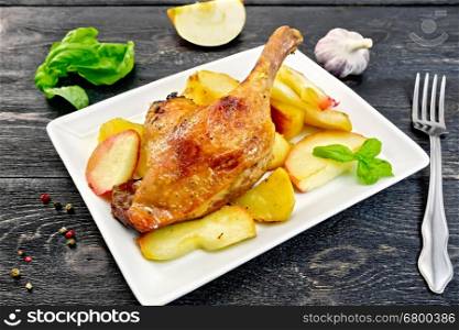 Roasted duck leg with apple, potatoes in a rectangular white plate, basil, garlic and fork on the background of wooden boards