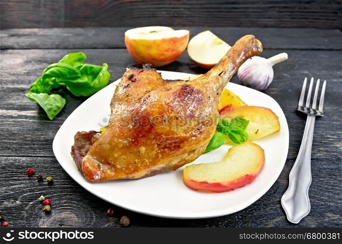 Roasted duck leg with apple, potatoes in a white plate, basil, garlic and fork on black background wooden plank