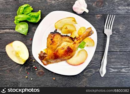 Roasted duck leg with apple, potatoes in a white plate, basil, garlic and fork on the background of the wooden planks on top