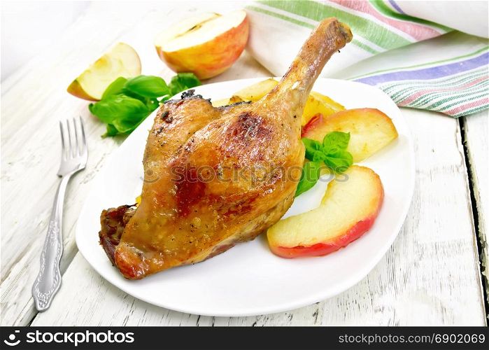 Roasted duck leg with apple, potatoes in a white plate, green basil, fork and kitchen towel on a background of wooden boards