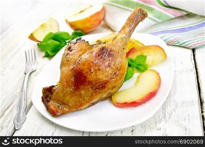 Roasted duck leg with apple, potatoes in a white plate, green basil, fork and kitchen towel on a background of wooden boards