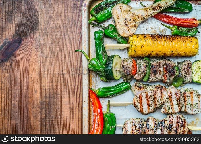 Roasted Meat skewers with vegetables and roasted corn ears on baking tray and rustic wooden background, top view, place for text. Meat food and grilling concept
