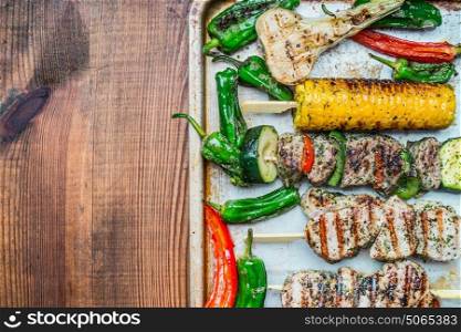 Roasted Meat skewers with vegetables and roasted corn ears on baking tray and rustic wooden background, top view, place for text. Meat food and grilling concept
