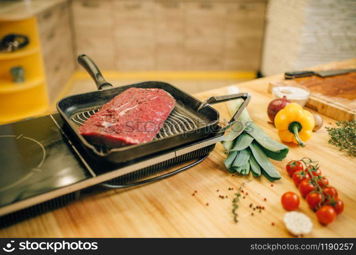 Roasted meat tenderloin in a frying pan on electric stove, vegetables on wooden board on background, nobody. Beef, seasonings and spices with herbs on countertop