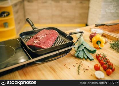 Roasted meat tenderloin in a frying pan on electric stove, vegetables on wooden board on background, nobody. Beef, seasonings and spices with herbs on countertop