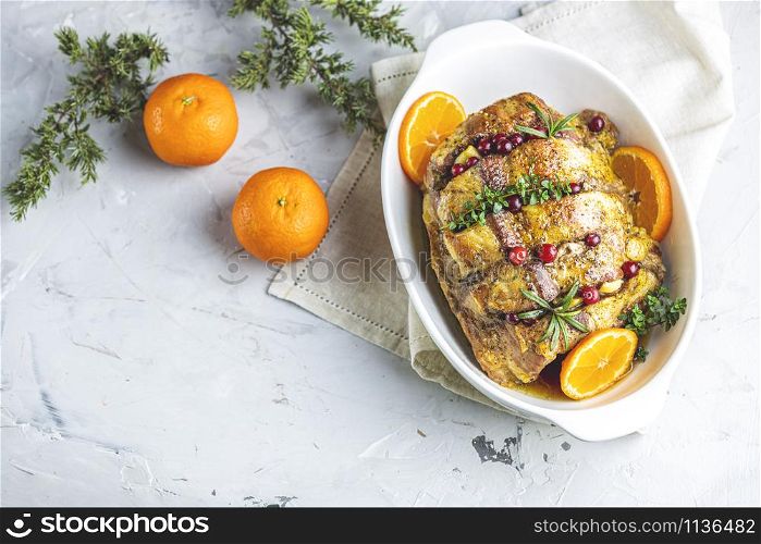 Roasted pork in white dish, christmas baked ham with cranberries, tangerines, thyme, rosemary, garlic on light table surface, close up.