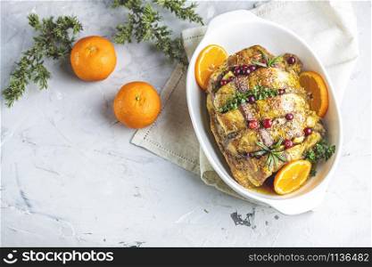 Roasted pork in white dish, christmas baked ham with cranberries, tangerines, thyme, rosemary, garlic on light table surface, close up.