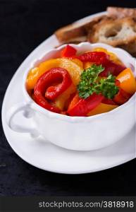 Roasted red and yellow peppers in white bowl with baguette over dark background, selective focus