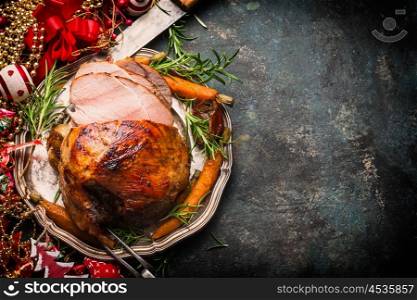 Roasted sliced Christmas ham on plate with fork, knife and festive decoration on dark rustic background, top view, border