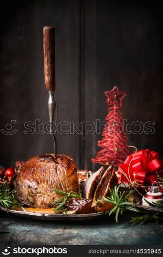 Roasted sliced Christmas ham with fork and red festive holiday decoration at dark wooden background, side view, place for text