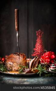 Roasted sliced Christmas ham with fork and red festive holiday decoration at dark wooden background, side view, place for text