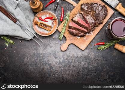 Roasted sliced grill steak on wooden cutting board with wine, seasoning and meat fork on dark vintage metal background, top view, border
