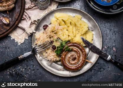 Roasted traditional sausage with potatoes and sauerkraut served on rustic table background with cutlery, top view