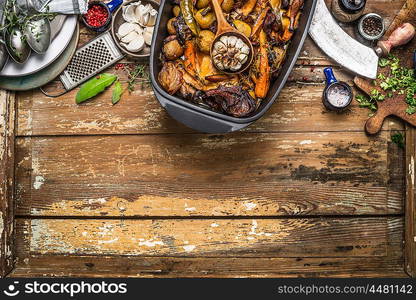 Roasted vegetables stew in cooking pot with kitchen tools on rustic wooden background, top view, border