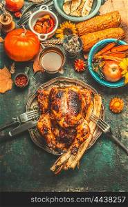 Roasted whole stuffed chicken or turkey for Thanksgiving Day dinner with sauce, pumpkins, corn and autumn harvest vegetables and cutlery on dark rustic background, top view